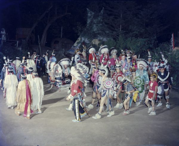 Slightly elevated view of a group of dancers in ceremonial clothing, performing the Green Corn dance. There is a teepee among trees in the background.