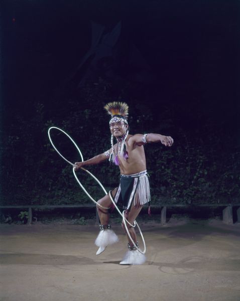 Hoop Dancer (Tony White Cloud), dressed in ceremonial clothing, is dancing with four hoops. Two hoops are in one hand, one of these two is also wrapped around his knee. The third hoop is around both legs, and the fourth is dangling on the opposite leg.