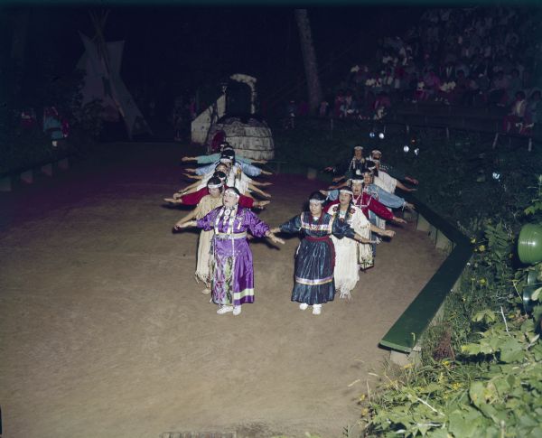 Elevated view of a group of women dressed in ceremonial clothing in two lines, with their arms out at the sides, performing the Swan Dance.