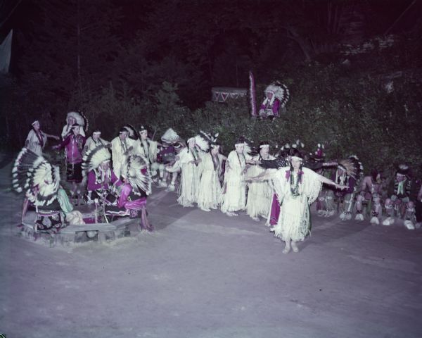 Women dressed in buckskins, performing the Wild Goose dance as men in headdresses watch.