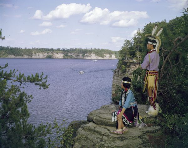 A man and women are posing on a rock formation and looking out over the big bay near Stand Rock. Boats are on the river in the distance.