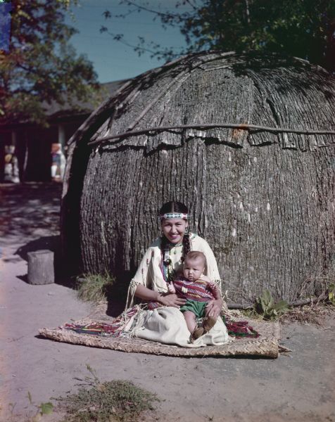 Portrait of a mother and child sitting on mat in front of a wigwam.  

"Bernodine Tallmadge and little son, Randy, in front of Elm Barl's wigwam."