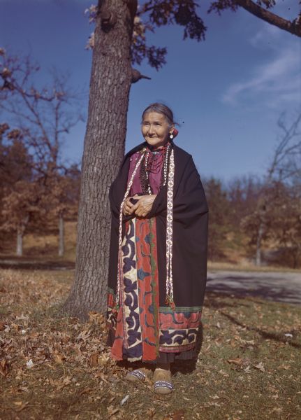 Full-length portrait of Suzie Redhorn wearing a black and red cloak. She is standing outside in front of a tree.