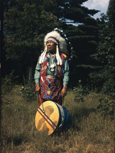 Outdoor full-length portrait of Chief Yellow Thunder. Trees are in the background, and he is looking off into the distance and is holding a drum which is resting on the ground.