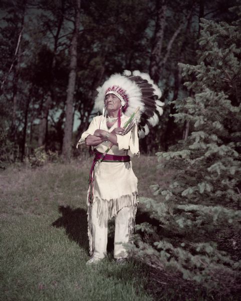 Full-length portrait of Little Pine Tree standing outside surrounded by trees. He is wearing buckskins and a headdress and has his arms crossed over his chest. 