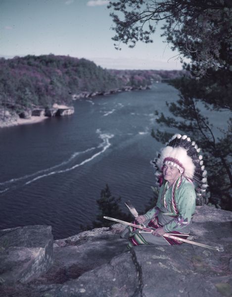 View towards a man wearing colorful clothing and a headdress, and holding a long bow and arrow. He is sitting on a rock formation high above the water.