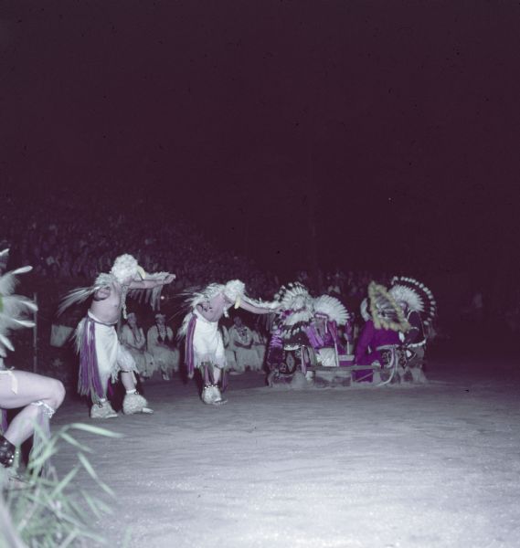 Two Native American Indian men dressed in eagle costumes are performing the Eagle Dance as an audience is watching in the background. 