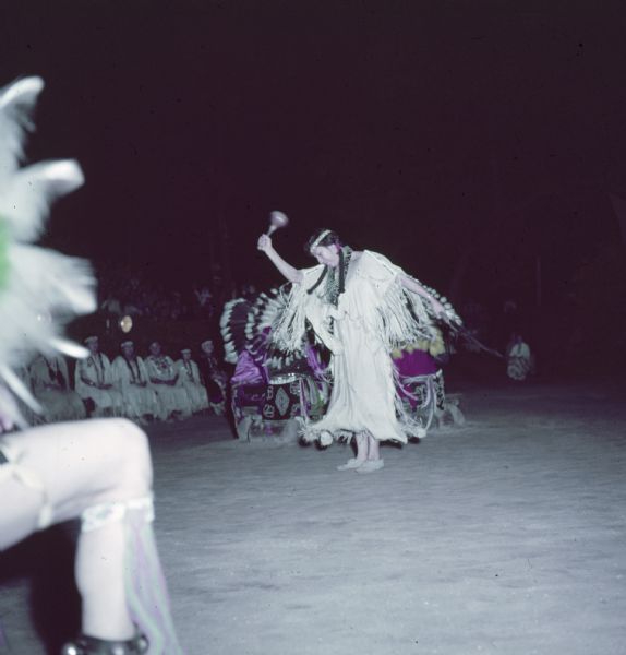 A Native American Indian women dressed in buckskin is performing the Swan Dance as an audience is watching from the left background.