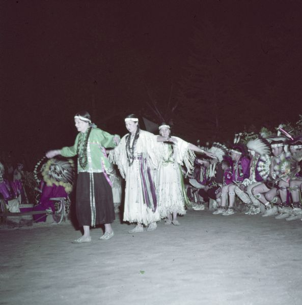 Three Native American Indian women are performing the Swan Dance at the Stand Rock Indian Ceremonial as an audience is watching in the background.