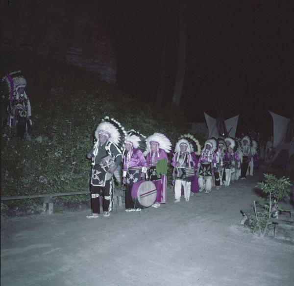 Native American Indian drummers, dressed in ceremonial clothing and headdresses, are lined up and walking up a path.