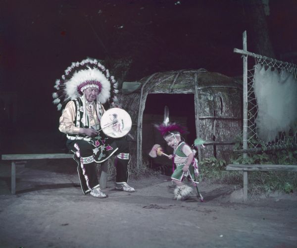 Blowsnake is sitting on a bench playing a drum, as little Bobby Bird is dancing. 