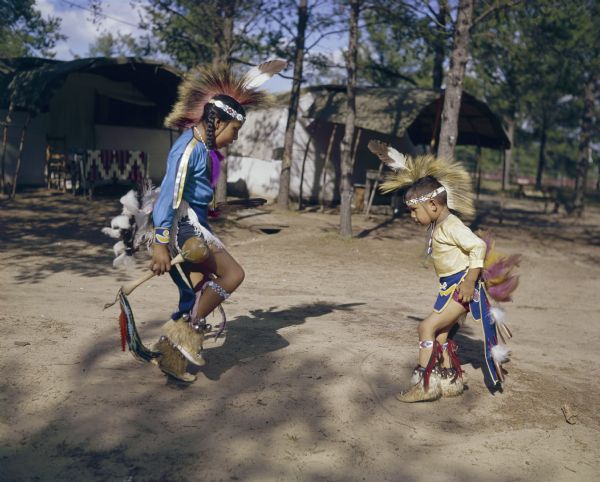 Two young Ho-Chunk children, both dressed in ceremonial clothing, are dancing outdoors at Pipe Dyer's Indian Village. The children are Janice (Lincoln) Rice, and her younger brother Jeff Lincoln.