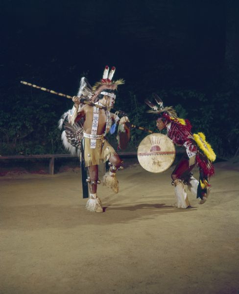 Two Native American Indian men, dressed in ceremonial clothing and holding spears and shields, are performing the Warrior's Dance at the Stand Rock Indian Ceremonial.