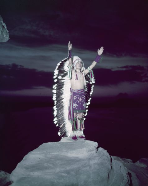 View towards a man wearing ceremonial clothing and a headdress standing on a rock formation with his arms in the air, performing the Sunrise Call of the Zuni.