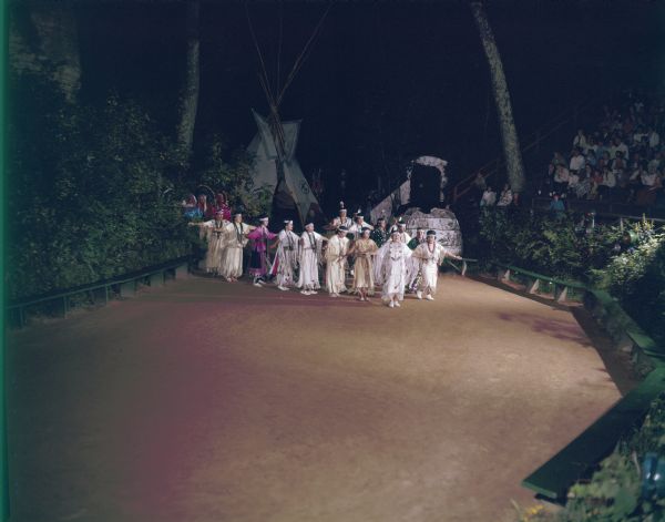 Elevated view of a group of women entering the stage while performing the Swan Dance at the Stand Rock Indian Ceremonial. An audience is watching while sitting on stands on the right.