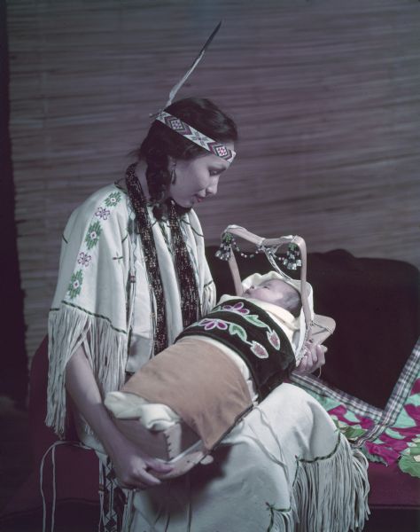 Portrait of a young woman in buckskins holding a baby in a cradle board.