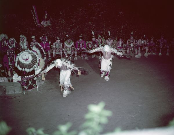 Slightly elevated view of two men performing the Eagle Dance, while an audience is watching from the side. The two men are dressed in eagle masks and feathered wings.
