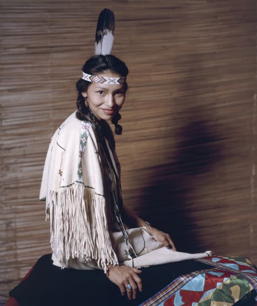 A portrait of an unidentified Native American Indian women dressed in a white buckskin dress. She is sitting on a quilt in front of a rush matted wall. 