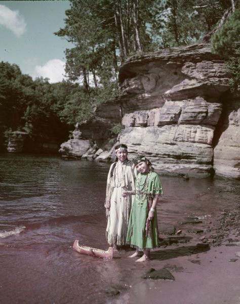 Two young women, Annette Miner and Carolyn Young, are standing in water on a sand bar with a toy canoe floating nearby.