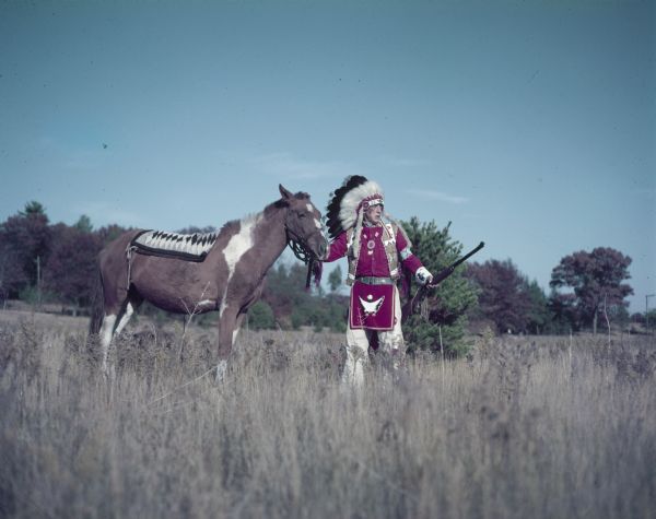 Roger Little Eagle (Tallmadge) standing in a field holding a horse by the bridle. 