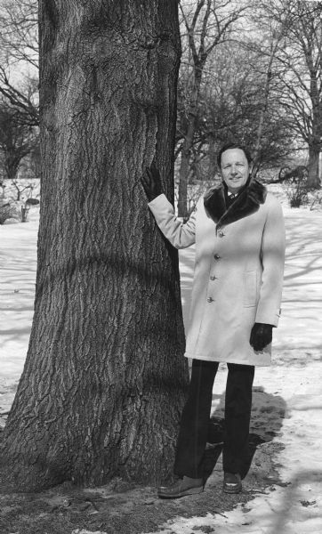Edward Hasselkus, University of Wisconsin professor of horticulture and curator of Longenecker Gardens at the University of Wisconsin Arboretum in Madison posing standing with his right hand resting on the trunk of a large tree. He is wearing a winter coat and gloves and there is snow on the ground. There is a cannon carriage and other trees in the background.