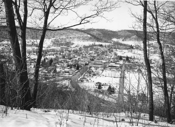 Bare trees frame an elevated view of Gays Mills taken from a snowy hilltop, showing Highway 131, lower right, as it curves into Main Street. Highway 171 enters Main Street diagonally from lower left. There are tall, snow-covered hills in the background, beyond the Kickapoo River, which is not clearly seen.  