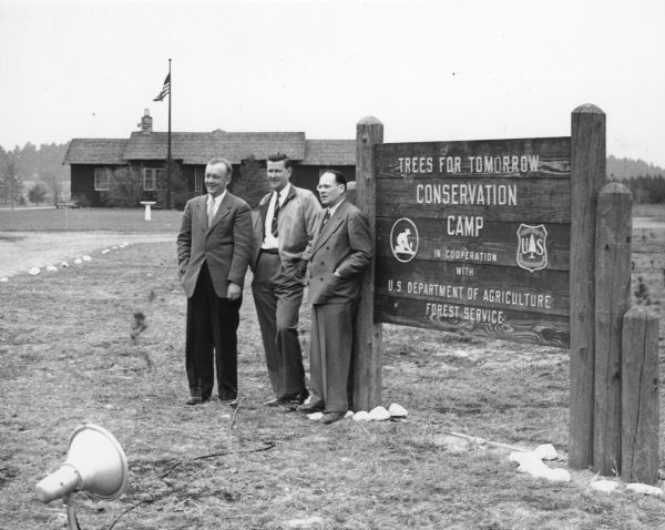 Forestors W.A. Sylvester and William Bjorge posing with Executive Director M.N. Taylor next to a sign at the Trees for Tomorrow Conservation Camp. There is a long, one-story building in the background with a flag pole and birdbath in front. Established in 1946, the camp is a private, nonprofit organization and accredited natural resources specialty school.