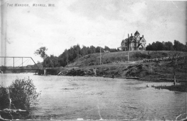 View across water towards a large Queen Anne style house sitting on a hill above the Wisconsin River. There is a bridge on the left and a road, now South Center Avenue, leads up the hill on the right.  