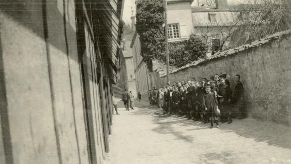 Students waiting outdoors during a sunny day, wearing their smocks and fatigue caps. Captioned: "The alley in which the school was located at Ste. Menehould. Cute youngsters with their smocks and 'fatigue' caps."