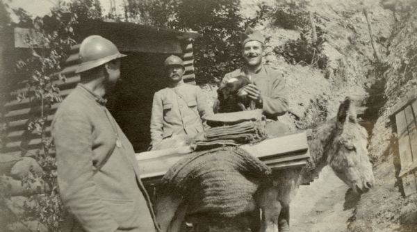 Three men loading a donkey with supplies. One of the men is holding a dog in his arms. Captioned: "Donkeys are used to carry munitions and supplies to batteries and in the trenches. They are a frightful bother when one is driving at night. Tour de Paris."  
