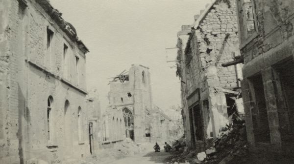Damaged buildings on the main road in Cormicy. Three men are in the road near a building on the right. Captioned: "The main street at Cormicy, showing the church in the distance. This road is on the way"