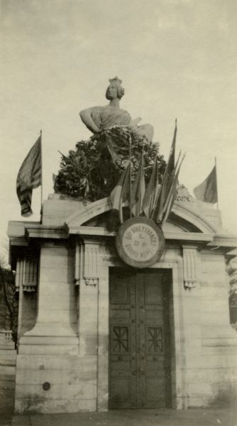 A monument decorated with flowers, flags and topped by a statue. Caption reads: "The Strassburg Monument of Alsace. Kept draped in mourning from 1871 to 1914, but since August 1914, it has been decorated with flowers and flags of the allies."
