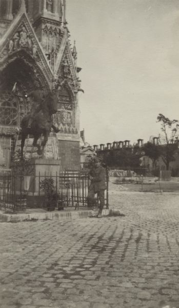 View of a man standing and posing near the statue of Jeanne d'Arc. Caption reads: "The most famous statue of Jeanne d'Arc, who visited Reims in the height of her fame to coronate Charles VII.
