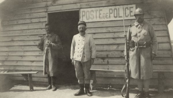 Three men standing guard in front of a building which is part of a prison camp in France. A sign on the building reads: "Post de Police." Caption reads: "Entrance to prison camp at St. Hilaire-au-Temple."