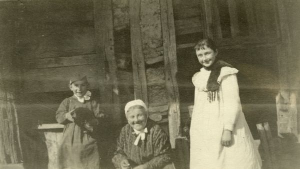 Two children and a woman posing near a building for a group portrait. Caption reads: "Waly, our first night stop out of Dombasle and where much exhilaration followed an animated Soccer game. The Boche were here for four days. Notice the typical French costumes."