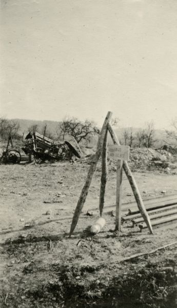 A non-exploded bomb is resting on the ground, over which three timbers have been placed. A sign reading "Obus Non Eclate" is on a post next to the bomb, and a wreckage of a vehicle is in the background. Caption reads: "A 77 'non eclate' which would have killed Sammy Lloyd had it exploded, but it didn't. St. Thomas." 