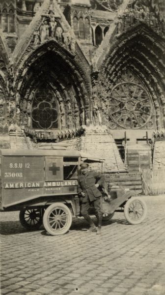 Portrait of a man standing and leaning against his ambulance, posing in front of a cathedral. Caption reads: "Reims. A close view of the entrance to the cathedral barred by an ambulance and ambulancier!" 