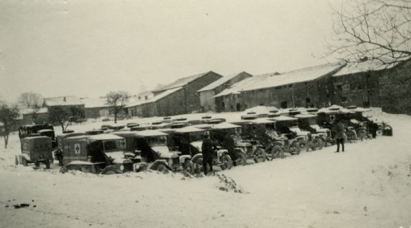 Two men standing in front of a large group of ambulances parked in the snow. Caption reads: "An early morning scene at Jubécourt. The drivers slept in their cars."