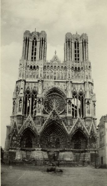 View of the front of the damaged Cathedral and the sculpture of Jeanne d'Arc in the front. Caption reads: "Reims. The front of the cathedral is scarcely damaged, but the interior is ruined by shells and fire. Notice the sand-bags around the bottom. Jeanne d'Arc, who coronated Charles VII, is in the foreground."