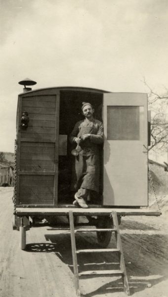 Portrait of a man posing at the open back door of his kitchenette, which is on the back of a truck. Caption reads: "Andre Gaillard, our 'bon cook' in the rear of his kitchenette. He was wounded at Revigny after killing the Boche who killed his brother, Dombasle."