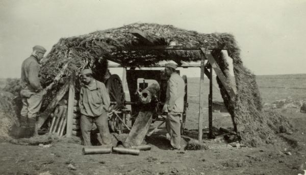 Group of men standing by a camouflaged gun base. Caption reads: "Famous French "75." Notice the empty shell, the loaded shells leaning against the sand-bags, and the grass cover to camouflage the gun from enemy aviators. Montgeville, Verdun sector."