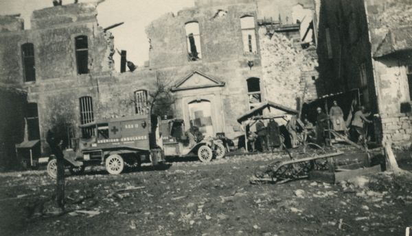 Several men standing near two ambulances which are parked near ruins. Caption reads: "The "Château," probably the most celebrated A.A.F.S poste. The entire cellar is utilized as operating rooms, dressing rooms and officers' quarters. Probably fifty men lived beneath the Château between Mort Homme and Hill 304."
