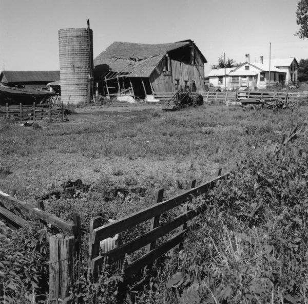 View over fence and field towards a dilapidated barn leaning against a silo. A tractor and a piece of agricultural machinery are parked in the field, and in the background a truck is parked near a farmhouse on the right.
