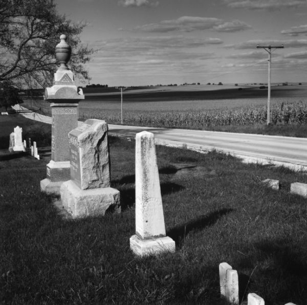 Gravestones and markers in a cemetery at the side of a paved road. A cornfield is across the road.