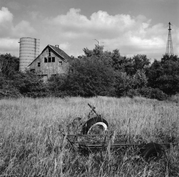 View across field towards an old barn and silo standing above a grove of trees and bushes. In the foreground is an old agricultural implement in the overgrown grass. A broken windmill is behind trees and a power line on the far right.