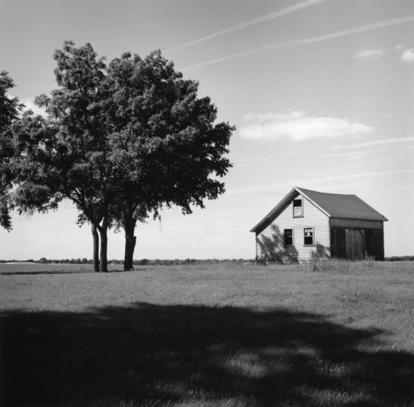 A small farm building standing in an open field of grass. Three trees are standing to the left of the building.