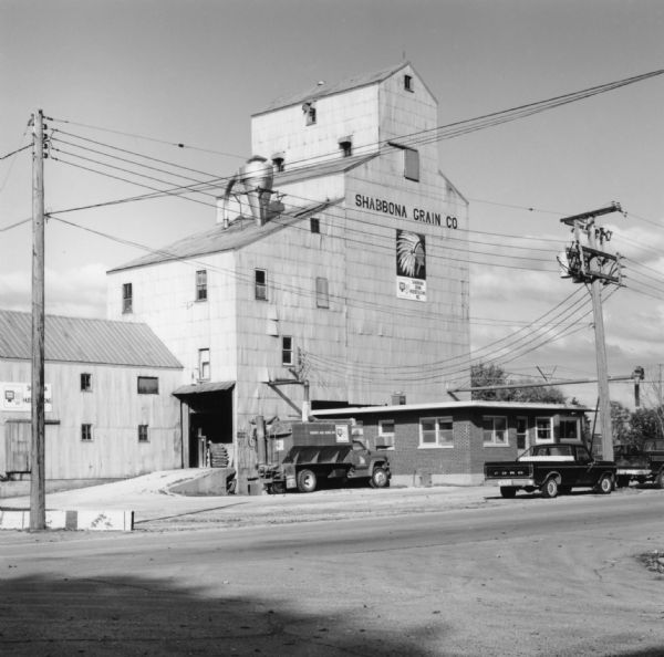 View across road towards trucks sitting in the parking lot in front of a large grain elevator. Grain is pouring out of a pipe into one of the loading trucks. Letters across the top of the elevator read "Shabbona Grain Co." Below them a sign displays an illustrated image of an American Indian Chief with a feathered headdress. This sign reads: "Shabbona Grain Hueber & Sons, Inc."