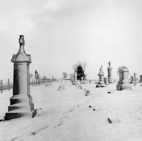 Gravestones in a cemetery standing in rows, their sides and the ground covered in snow. A rope or chain fence separates the cemetery from the road along the left. A set of footprints is leading from the right diagonally down to the lower left corner. A few trees are on the horizon.