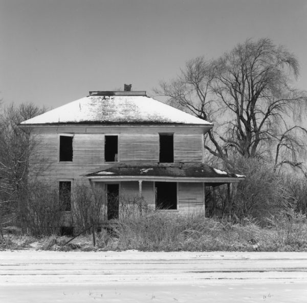 An abandoned house surrounded by uncut grass and trees on the side of a road. Snow is covering the ground and lightly covers the roof of the two-story house. The windows of the house are missing.