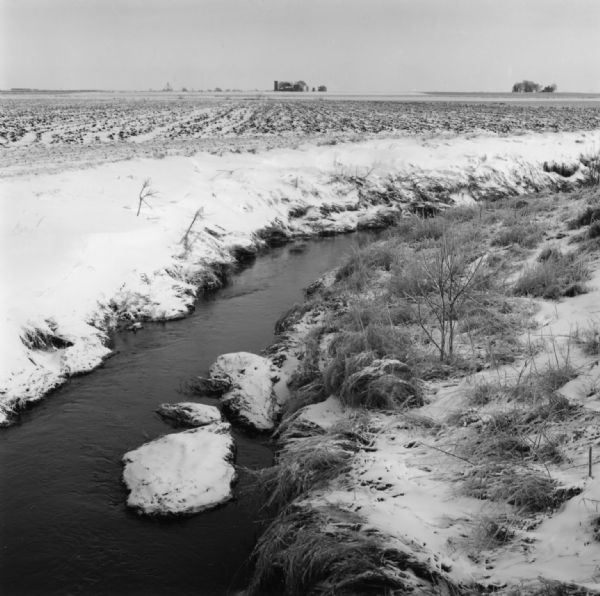 A small river winding through a farm field, with snow covering the banks and field. Trees and a house are on the horizon.